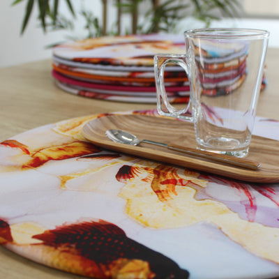 Bajoplato Funda Pez sobre mesa de madera junto a una pila de mantelitos de colores y taza de cristal